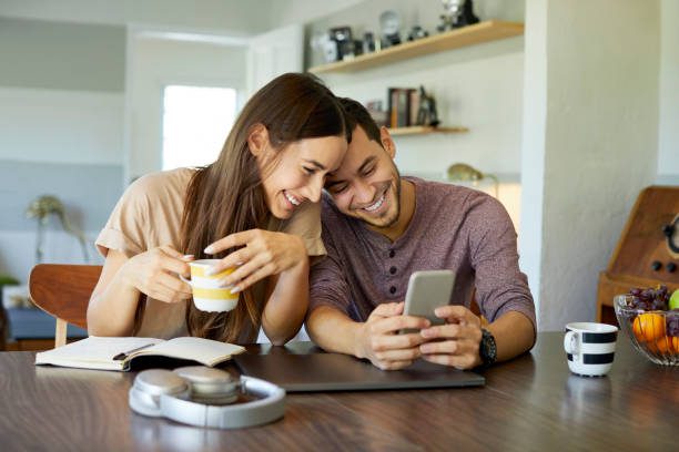 istockphoto-1133527315-612×612 Cheerful boyfriend showing mobile phone to girlfriend in dining room. Young woman is holding coffee cup. They are spending leisure time together at home.